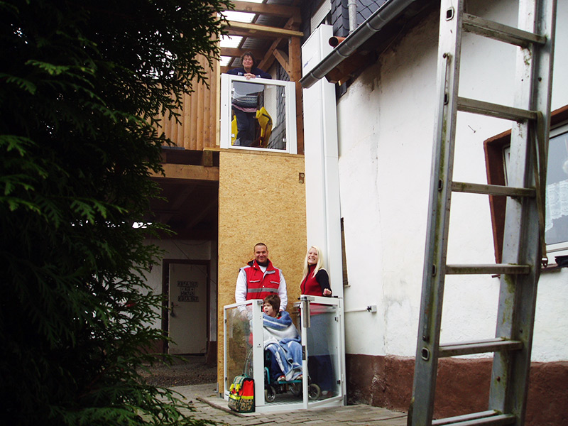 Durch Spenden der AfbM Hessen e.V., der AfbM Waldeck - Frankenberg e.V. und anderer Vereinigungen konnte ein Außenfahrstuhl an einem Haus finanziert werden, um der Mutter eines Jungen mit einer schweren Behinderung das Tragen über die Treppe zu ersparen. Die Familie vor dem Außenfahrstuhl